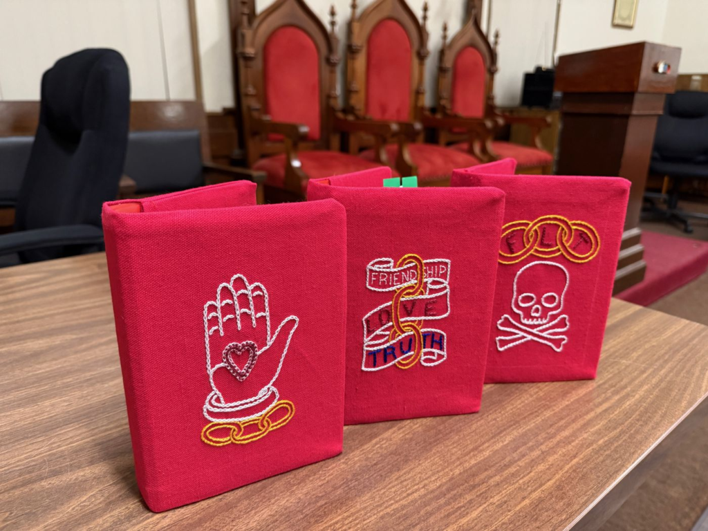 Three red fabric charge book covers embroidered with Odd Fellows symbols: a hand holding a heart, the three links with a Friendship, Love, Truth banner, and the FLT links over a skull and crossbones