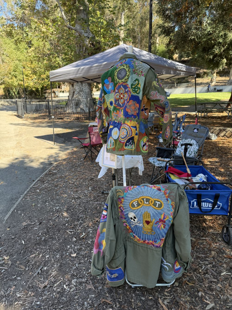 Two olive green jackets covered in colorful hand embroidery, one with the Odd Fellows three links, a hand with a heart, and a skull and crossbones on the back